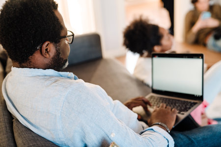 African american man using laptop while sitting on sofa with family at homeの写真素材