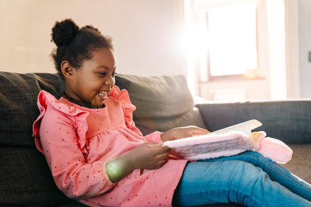 African american little girl sitting on sofa and reading her notebook at homeの写真素材