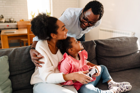 African american family with little daughter sitting on sofa and playing video game at homeの写真素材