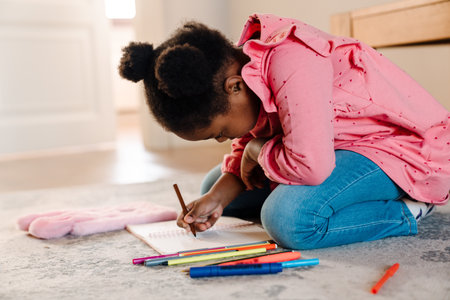 African american little girl sitting on floor and drawing with markers at home