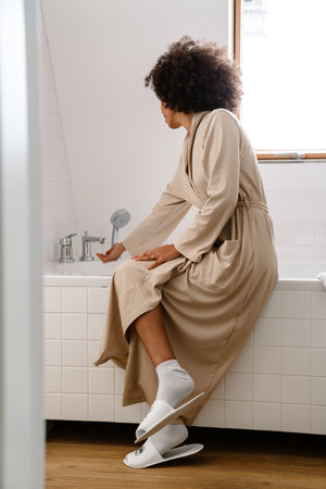 African american young woman with curly hairstyle filling bathtub with water in morningの写真素材