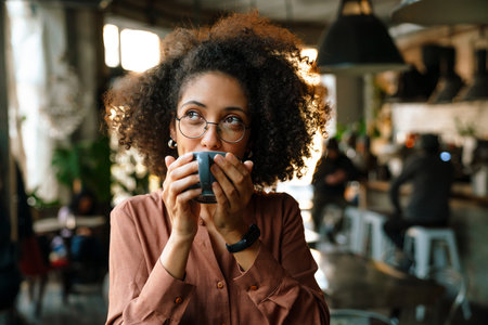 Young african american woman wearing eyeglasses drinking coffee in cafeの写真素材
