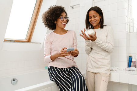 Happy african american woman and her teenage daughter doing beauty skincare in bathroomの写真素材