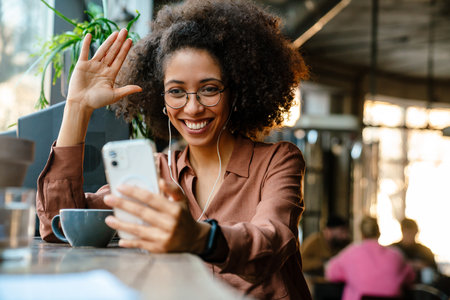 Young black woman with afro hairstyle using cellphone and earphones in cafe indoorsの写真素材