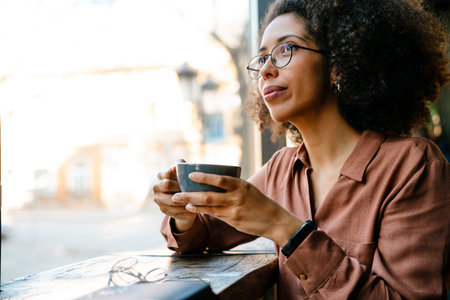 Young black woman with afro hairstyle smiling and drinking coffee in cafe indoorsの写真素材