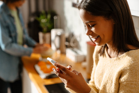 Black girl using mobile phone during spending time with her mother in kitchen at homeの写真素材