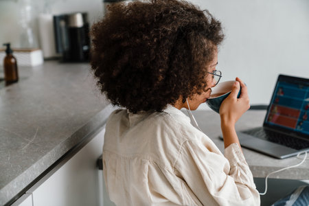 Black woman in earphones drinking coffee while working with laptop at homeの写真素材