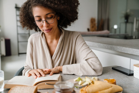 Black woman wearing eyeglasses reading book while having breakfast at homeの写真素材