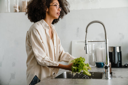 Black woman wearing eyeglasses washing lettuce while cooking at homeの写真素材