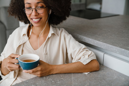 Black woman in earphones listening music while drinking tea at homeの写真素材