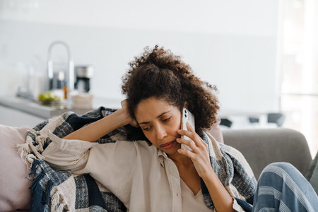 Black displeased woman talking on mobile phone while sitting on sofa at homeの写真素材