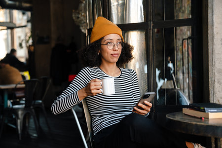 Young african american woman drinking tea and using cellphone in cafeの写真素材