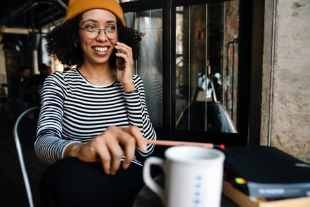 Young african american woman smiling while talking on cellphone in cafeの写真素材