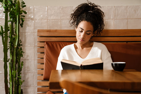 Young black woman drinking coffee and reading book in cafe indoorsの写真素材