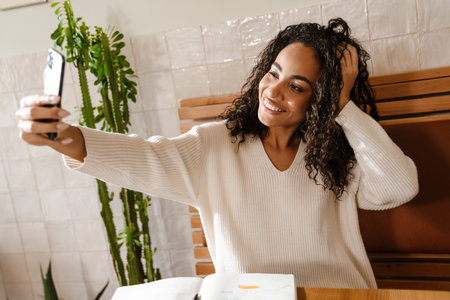 Young black woman smiling and taking selfie photo on cellphone in cafe indoorsの写真素材