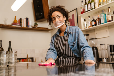 Black barista woman wearing face mask smiling while working in cafe indoorsの写真素材