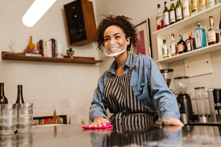 Black barista woman wearing face mask smiling while working in cafe indoorsの写真素材