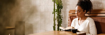 Young black woman drinking coffee and reading book in cafe indoorsの写真素材