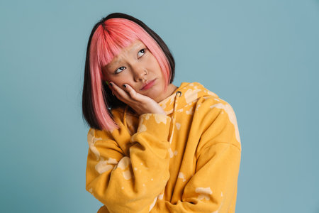 Asian girl with pink hair and piercing frowning while looking aside isolated over blue background