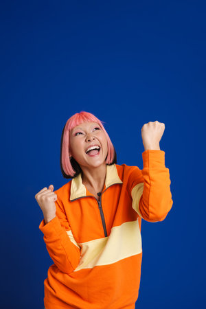 Asian girl with pink hair screaming while making winner gesture isolated over blue background