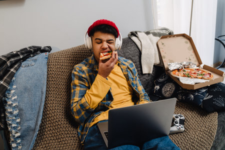 Middle-eastern teenage boy eating pizza and using laptop while sitting on sofa at home