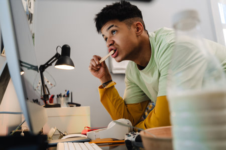 Brunette teenage boy brushing his teeth and looking at desktop computer in bedroom