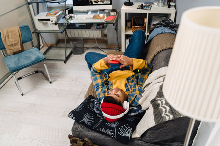 Middle-eastern teenage boy using headphones and cellphone while lying on couch at home