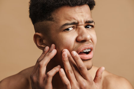 Young shirtless man frowning while squeezing pimple on his face isolated over beige background