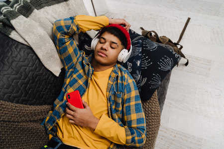 Middle-eastern teenage boy using headphones and cellphone while lying on couch at homeの写真素材