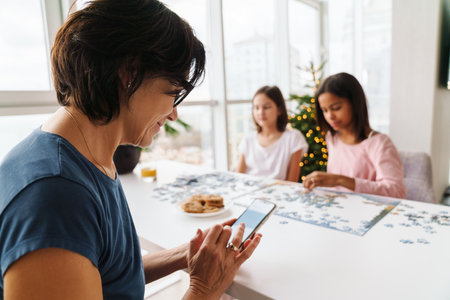 Multiracial sisters doing puzzle together while their mother using cellphone at homeの写真素材