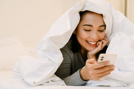 Young smiling asian girl in home suit lying with phone on bed under covers at homeの写真素材