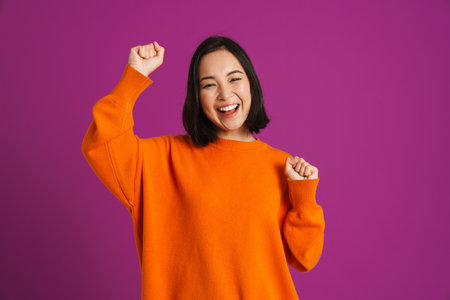 Young asian woman laughing while making winner gesture isolated over purple backgroundの写真素材