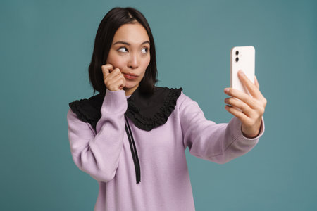 Young asian woman looking aside while taking selfie on cellphone isolated over blue background