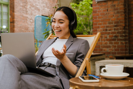 Asian young woman in headphones working with laptop while sitting in cafe outdoors