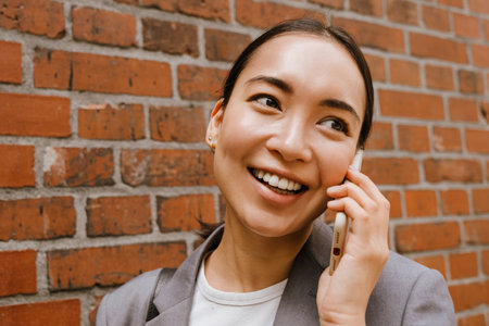 Asian young businesswoman smiling and talking on cellphone while standing by wall on streetの写真素材