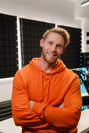 Bearded white young man standing by radio workplace while broadcasting in studio