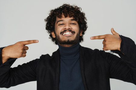 Young handsome curly smiling indian man in black coat pointing on himself over isolated grey backgroundの写真素材