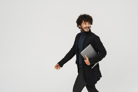 Young handsome curly smiling indian man walking with laptop over isolated grey backgroundの写真素材