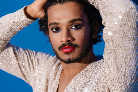 Young curly man with makeup posing and looking at camera isolated over blue background