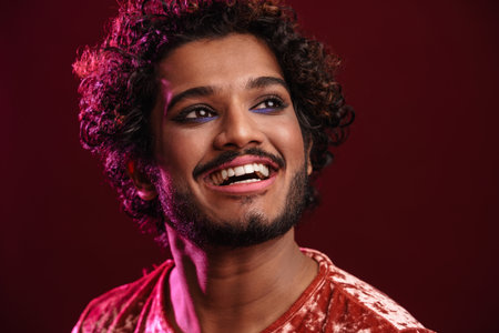 Young curly man with makeup laughing and looking aside isolated over red backgroundの写真素材
