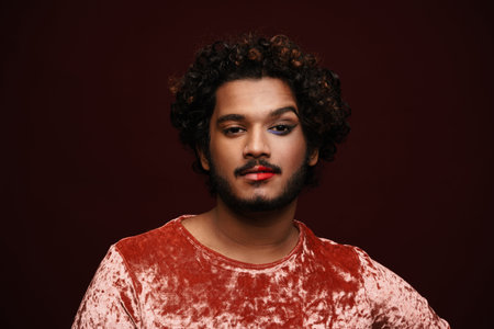 Young curly man with makeup posing and looking at camera isolated over red background
