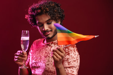 Young man with rainbow flag drinking champagne and smiling isolated over red background