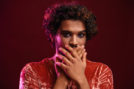 Young scared man with makeup covering his mouth and looking camera isolatedの写真素材