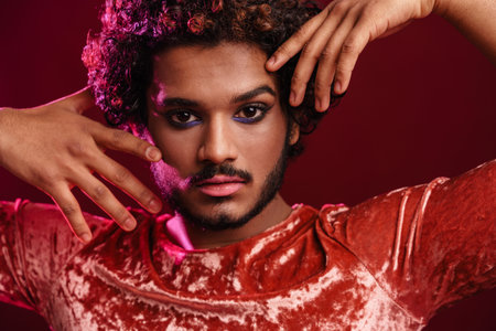 Young curly man with makeup gesturing and looking at camera isolated over red background