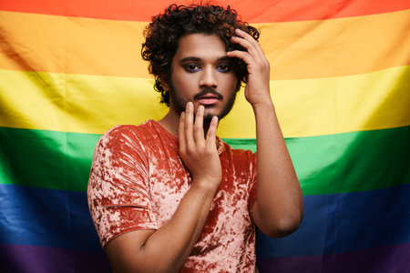 Young curly man with makeup posing and looking at camera isolated over rainbow backgroundの写真素材