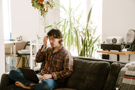 White man wearing shirt using laptop while resting on couch at homeの写真素材