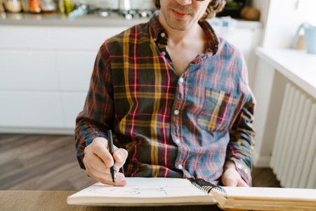 White man wearing glasses writing down notes while sitting in kitchen at home