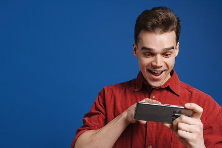 Excited white man playing online game on cellphone isolated over blue background