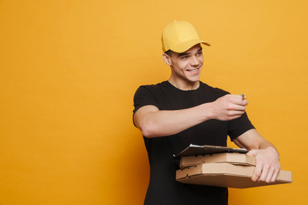Young delivery man smiling while posing with clipboard and pizza boxes isolated over yellow...