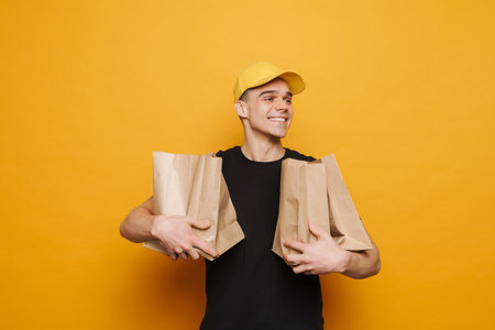 Young delivery man smiling while posing with paper bags isolated over yellow background